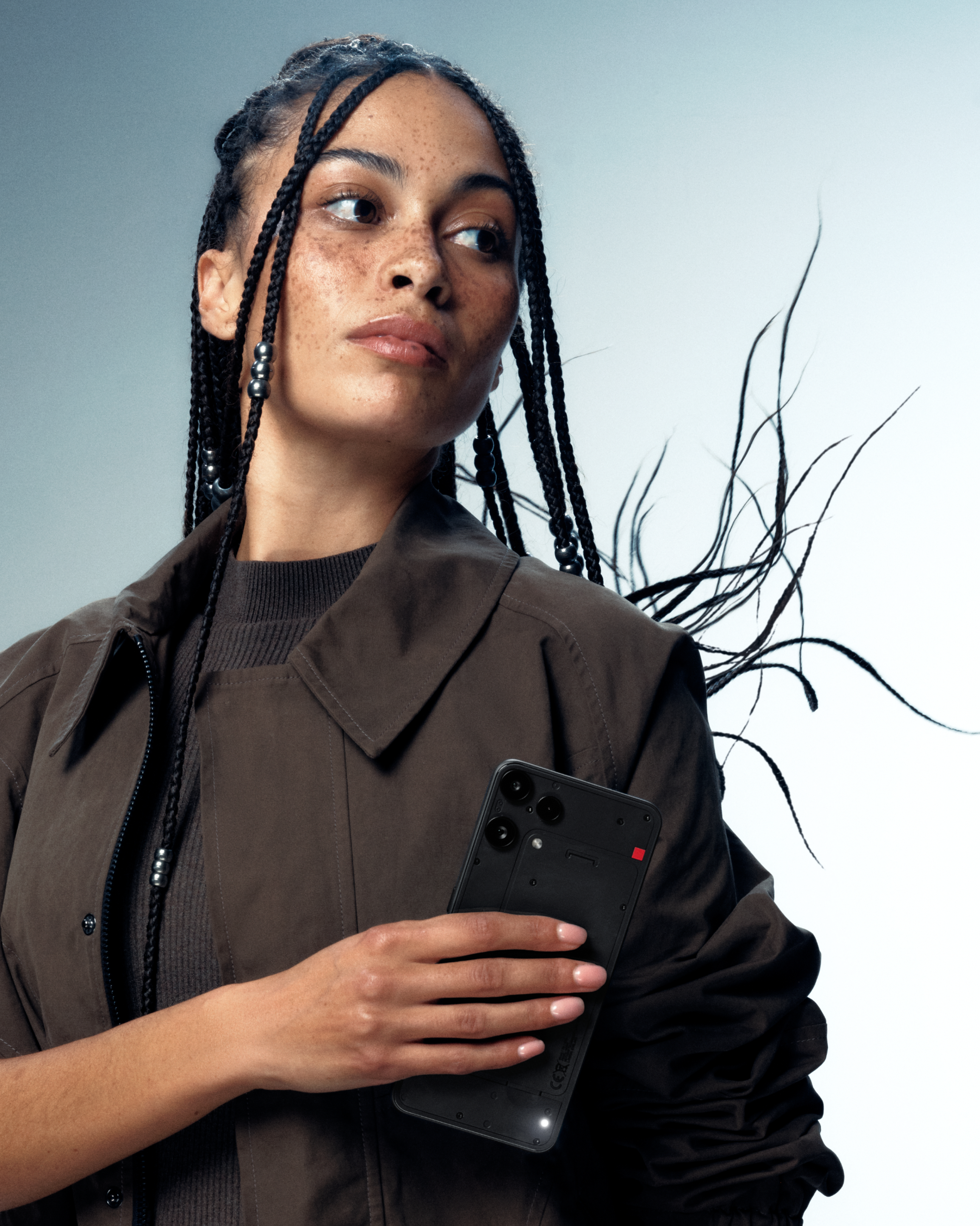 Eine Frau mit Zöpfen hält ein schwarzes Telefon. Sie trägt eine braune Jacke. Der Hintergrund ist hellgrau.